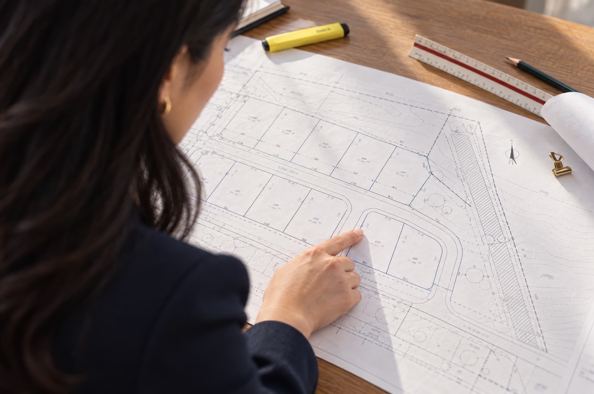 Site plans and a subdivision layout spread across a table, seen over Christine's shoulder